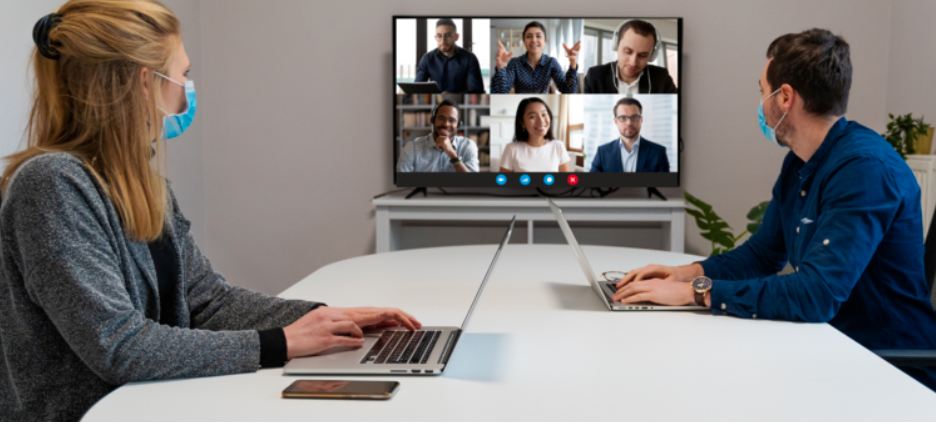 A hybrid team meeting, two members in person with masks, 6 team members on video conference. 