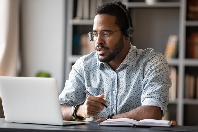 Man working virtually on a laptop, writing notes in a notebook.